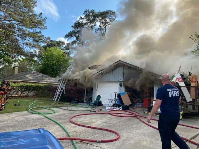 A firefighter in uniform stands near a house with smoke billowing from the roof. A ladder leans against the house, and fire hoses are scattered on the ground. Another person in firefighting gear is visible to the left amid trees and bushes.