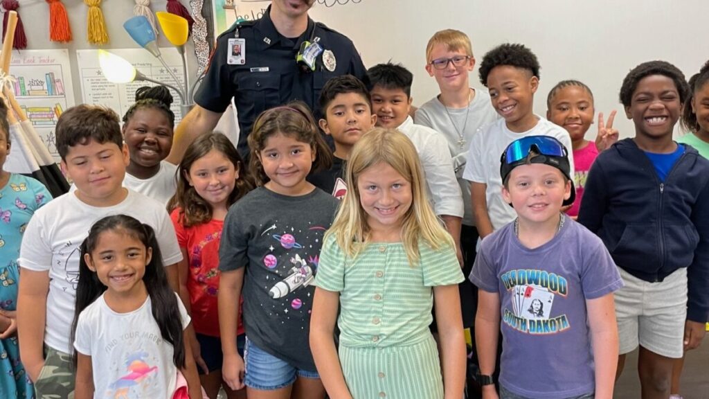 A group of smiling children and a police officer pose together in a classroom. The officer stands in the middle, surrounded by the children, who are of diverse ethnicities. Classroom decorations are visible in the background.