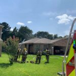 Firefighters in gear respond to a smoke-emitting house in a suburban neighborhood. A fire truck is partially visible on the right. A group of trees and a clear sky are in the background.