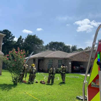 Firefighters in gear respond to a smoke-emitting house in a suburban neighborhood. A fire truck is partially visible on the right. A group of trees and a clear sky are in the background.