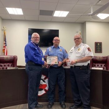 Three men standing in a conference room. The man in the middle is holding a certificate. The two others are shaking his hands. There is an American flag and a large screen in the background.