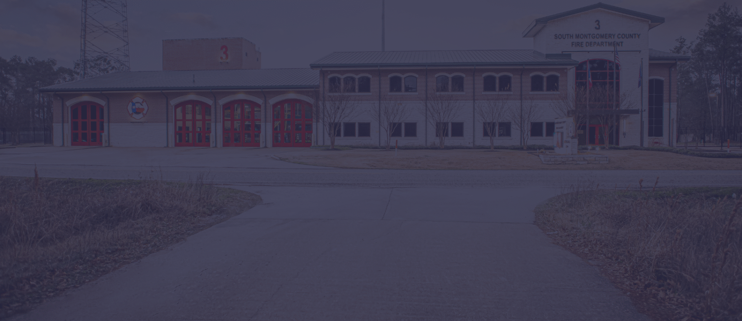 A two-story fire station with red garage doors and a sign reading South Montgomery County Fire Department above the main entrance. The building is surrounded by trees and a wide driveway.