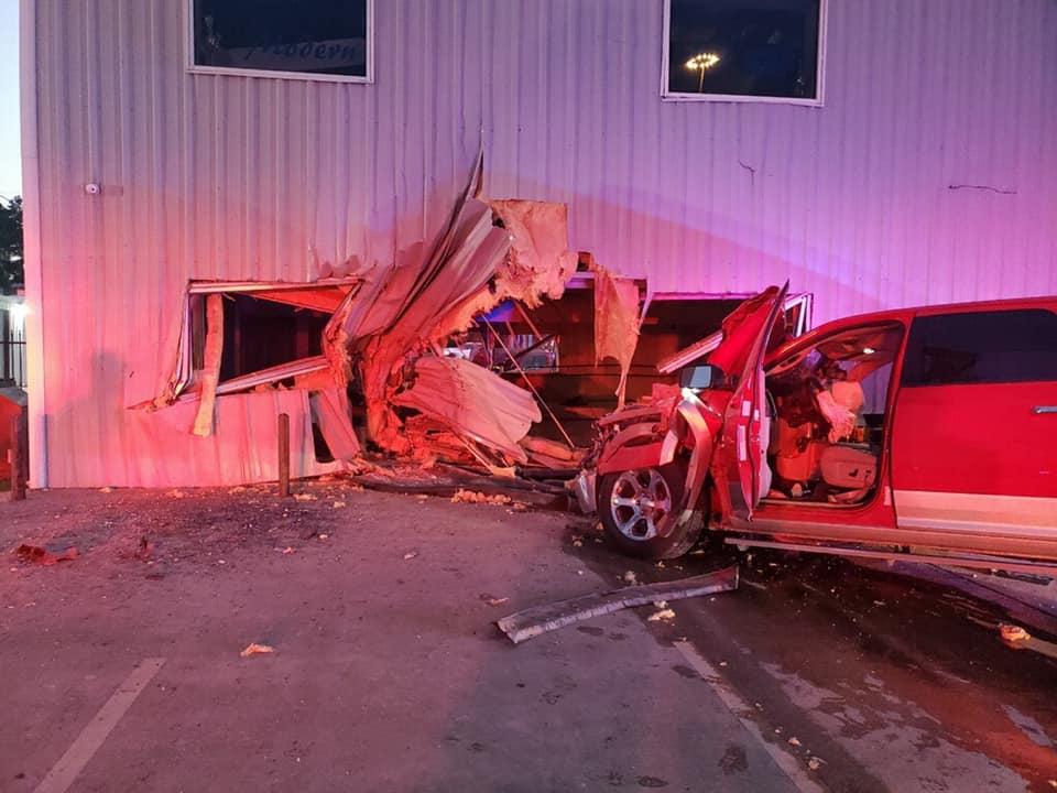 A red pickup truck has crashed into the side of a building, creating a large hole. The exterior wall is heavily damaged with debris scattered around. The truck is partially inside the building, with its front end barely visible.
