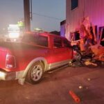 A red pickup truck has crashed into the corner of a building, causing significant structural damage. Debris and wood are scattered around the scene. The rear of the truck is slightly elevated.