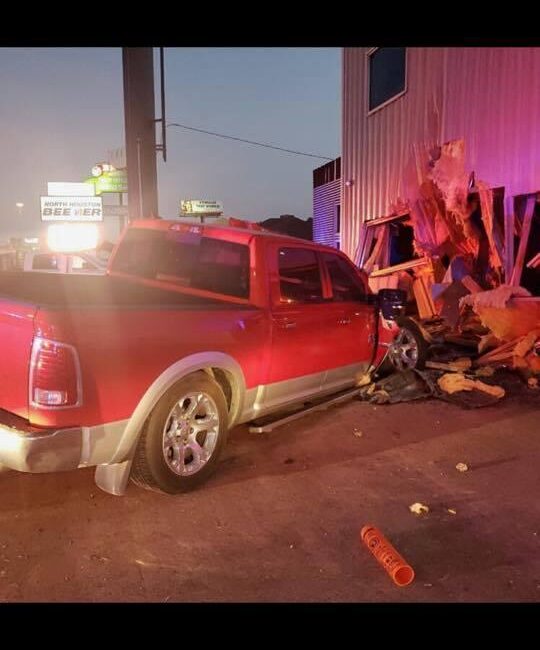 A red pickup truck has crashed into the corner of a building, causing significant structural damage. Debris and wood are scattered around the scene. The rear of the truck is slightly elevated.