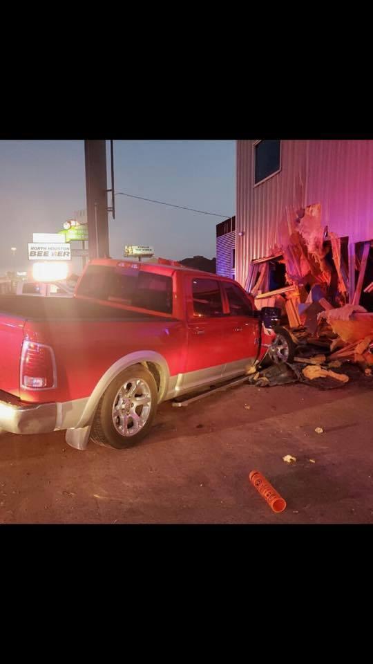 A red pickup truck has crashed into the corner of a building, causing significant structural damage. Debris and wood are scattered around the scene. The rear of the truck is slightly elevated.