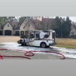 A burnt-out delivery truck with a charred front and foam residue around stands on a suburban street, surrounded by hoses. Large houses and trees are in the background.