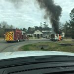 A firetruck and firefighters respond to a burning vehicle on a residential street. Thick black smoke rises from the vehicle, surrounded by trees and a large white house in the background. Emergency responders are preparing to extinguish the fire.