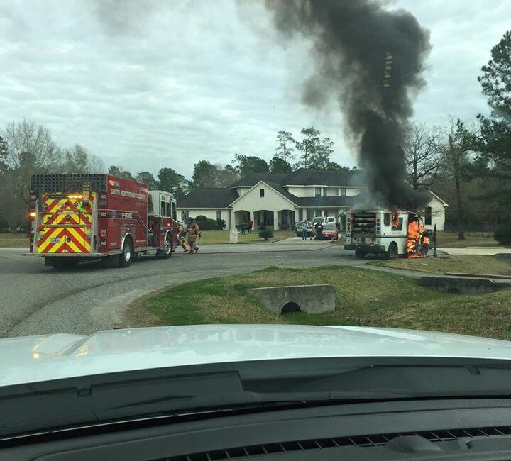 A firetruck and firefighters respond to a burning vehicle on a residential street. Thick black smoke rises from the vehicle, surrounded by trees and a large white house in the background. Emergency responders are preparing to extinguish the fire.