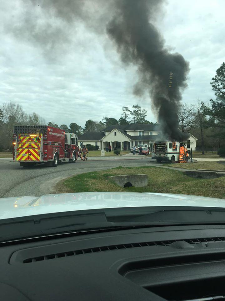 A firetruck and firefighters respond to a burning vehicle on a residential street. Thick black smoke rises from the vehicle, surrounded by trees and a large white house in the background. Emergency responders are preparing to extinguish the fire.