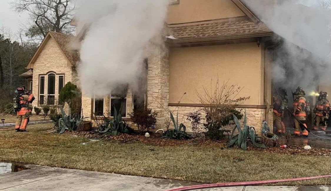 Firefighters in gear respond to a house fire with smoke billowing from the roof and windows. Hoses are laid out on the ground in front of the house, while greenery surrounds the property.