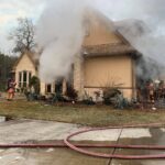 Firefighters in gear respond to a house fire with smoke billowing from the roof and windows. Hoses are laid out on the ground in front of the house, while greenery surrounds the property.