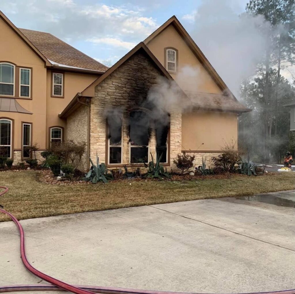 A two-story house with a charred exterior around the front windows shows smoke billowing out. Fire hoses are on the driveway, and a firefighter can be seen in the background, indicating a recent fire.
