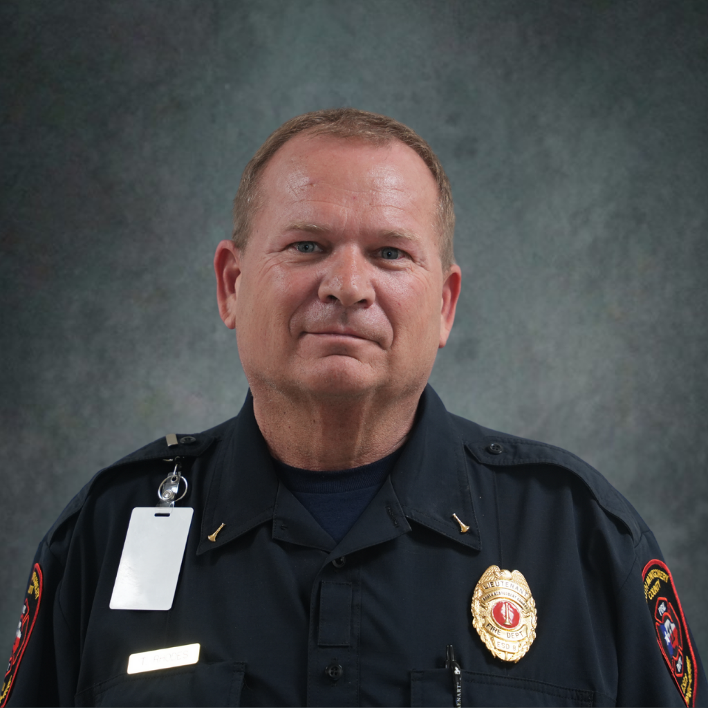 A middle-aged man in a dark fire department uniform with badges and shoulder patches is standing in front of a grey textured background, facing the camera and smiling slightly.