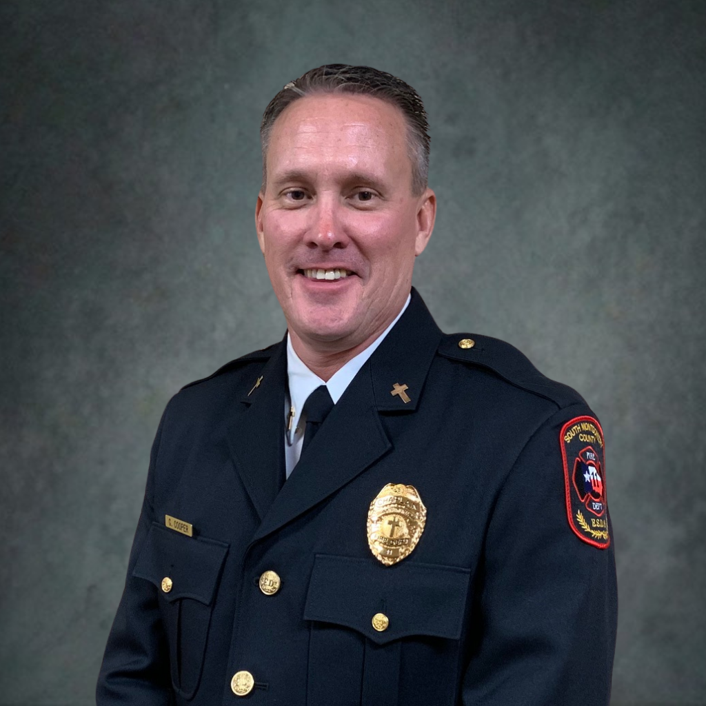 A man in a dark formal fire department uniform with badges and a patch, smiling in front of a plain, textured gray background.
