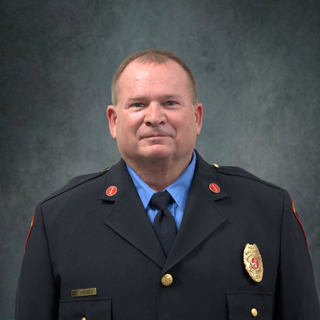 A middle-aged man in a formal dark firefighter uniform poses against a gray textured background. He wears a blue shirt, dark tie, gold badge, red collar pins, and a nameplate reading M. VARADY.