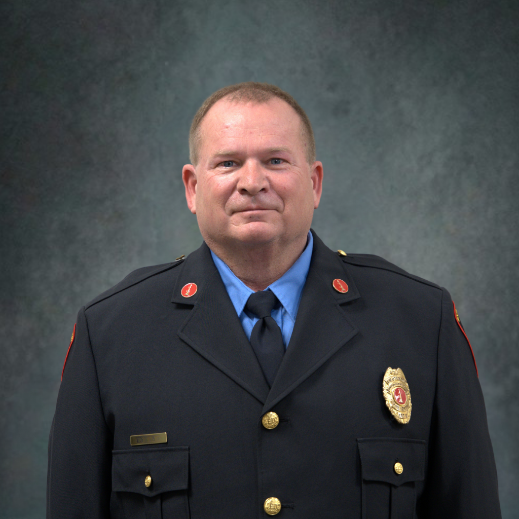 A middle-aged man wearing a formal firefighter’s dress uniform with a badge, pins, and a tie stands against a mottled gray background, looking directly at the camera with a neutral expression.
