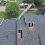 A fire truck with an extended ladder is parked near a building with two large burnt holes in the roof, surrounded by trees and a grassy area.