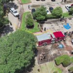 Aerial view of firefighters and several fire trucks in a parking lot, with hoses on the ground and people gathered nearby, some shaded by trees and others standing around emergency vehicles.