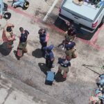 Aerial view of firefighters in gear standing near a white truck in a parking lot, with equipment scattered nearby and a red fire truck partially visible.