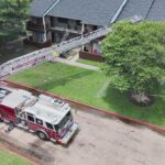 A fire truck with an extended ladder is parked near an apartment building. The ladder reaches up to a balcony, and caution tape surrounds the area. Trees and grassy lawns are visible.
