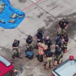 A group of firefighters in gear stands together in a parking lot near a red fire truck, with air tanks and equipment laid out on a blue tarp and the ground nearby.