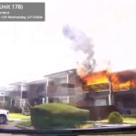 An apartment building is on fire, with flames and smoke coming from the upper floor. Emergency vehicle details and timestamp are displayed in the top left corner.