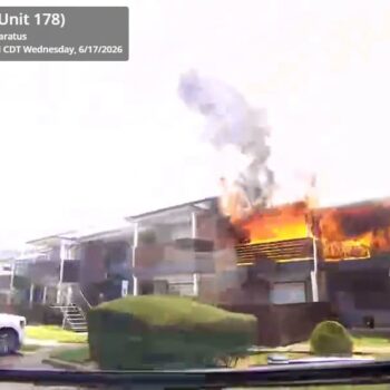 An apartment building is on fire, with flames and smoke coming from the upper floor. Emergency vehicle details and timestamp are displayed in the top left corner.