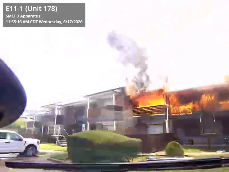 An apartment building is on fire, with flames and smoke coming from the upper floor. Emergency vehicle details and timestamp are displayed in the top left corner.