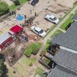 Aerial view of firefighters, police, and bystanders near a fire truck and police SUV in an apartment parking lot. Hoses are laid out and water is sprayed as part of an emergency response.