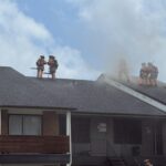Four firefighters in gear stand on a smoky roof of a building, working to ventilate the smoke. Two are holding tools while two others kneel and cut into the roof, as smoke billows around them.