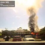 A large fire with heavy black smoke rises from a building behind parked cars under a clear sky, as seen from a vehicle dashboard camera.