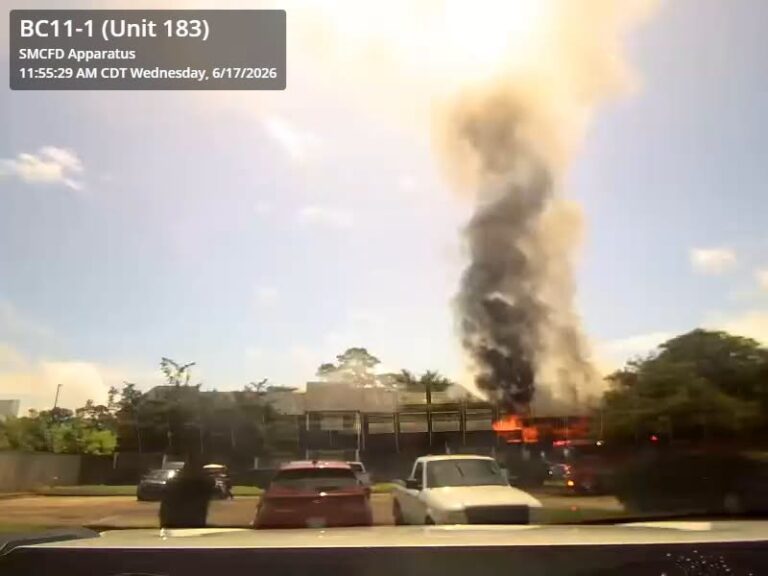 A large fire with heavy black smoke rises from a building behind parked cars under a clear sky, as seen from a vehicle dashboard camera.