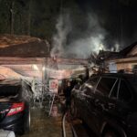 Smoke rises from a burning house at night, with firefighters responding. Several cars are parked nearby, and hoses run along the wet ground as light from the fire and emergency lights illuminates the scene.