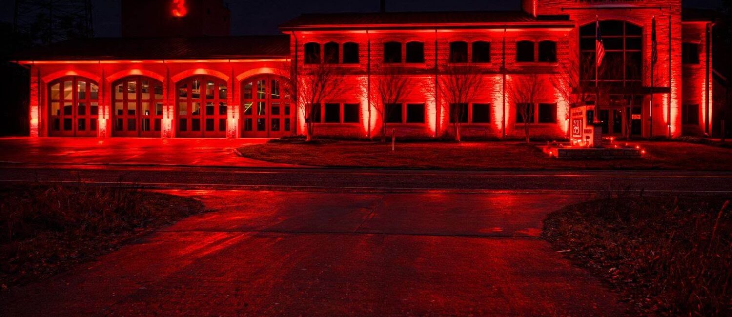 A fire station building illuminated by bright red lights at night. The number 3 is visible on the structure, and bare trees line the front. The scene reflects on the wet pavement.