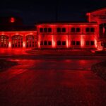A fire station building illuminated by bright red lights at night. The number 3 is visible on the structure, and bare trees line the front. The scene reflects on the wet pavement.