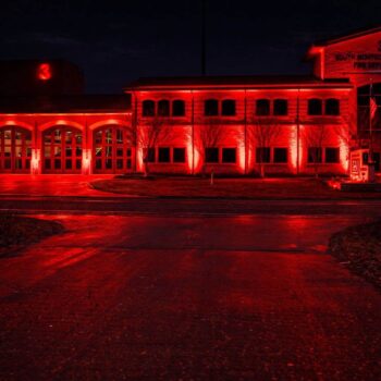 A fire station building illuminated by bright red lights at night. The number 3 is visible on the structure, and bare trees line the front. The scene reflects on the wet pavement.