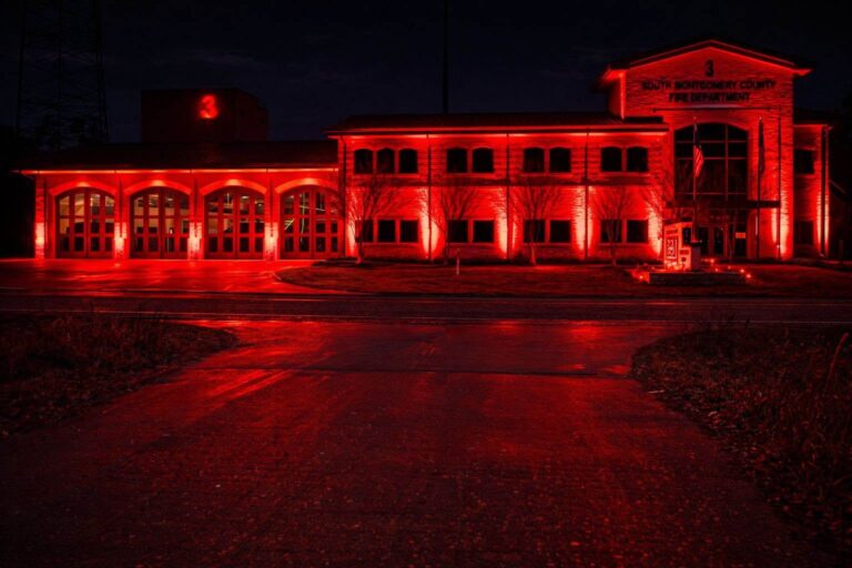 A fire station building illuminated by bright red lights at night. The number 3 is visible on the structure, and bare trees line the front. The scene reflects on the wet pavement.