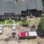 Aerial view of firefighters and a fire truck in front of an apartment building with visible fire damage on the roof. Ladders lean against the building, and hoses are spread across the ground.