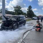 A firefighter sprays foam on a severely burned SUV on a road, with police cars, fire truck, and other people in the background under a partly cloudy sky.