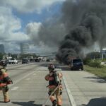 Two firefighters approach a burning SUV on the highway, dragging a hose. Thick black smoke rises from the vehicle as cars pass by in surrounding lanes under a partly cloudy sky.