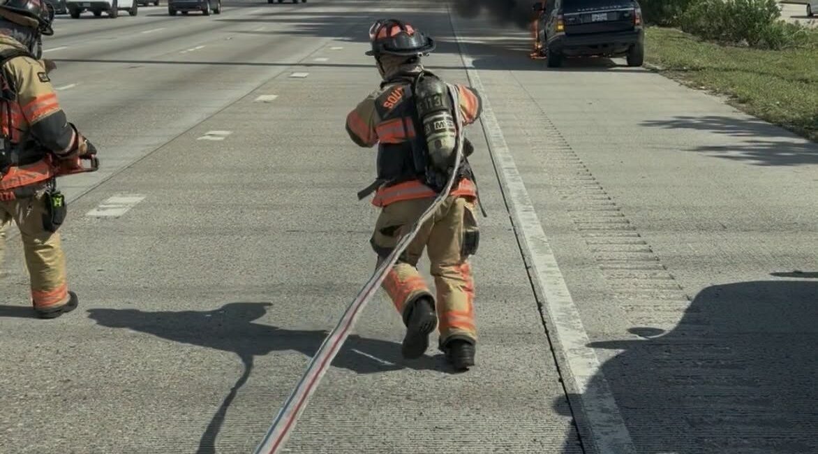 Two firefighters in gear run toward a smoking, burning vehicle on the side of a busy highway, pulling a fire hose while dark smoke rises into the cloudy sky.