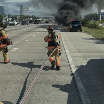 Two firefighters in gear run toward a smoking, burning vehicle on the side of a busy highway, pulling a fire hose while dark smoke rises into the cloudy sky.
