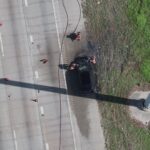 Aerial view of firefighters inspecting a burned car on the side of a highway near a grassy area, with hoses on the road and a large shadow from a nearby structure.