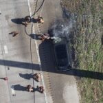 Aerial view of firefighters surrounding a black car on a road, spraying water on its hood; water and debris are visible nearby.