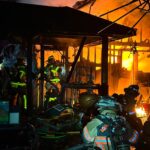 Firefighters in full gear battle intense flames inside a heavily damaged, burning building at night. Debris is scattered on the ground as the fire glows brightly in the background.