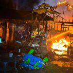 Firefighters work to extinguish a burning building at night. Flames and smoke rise, with charred debris and a blue and green childrens toy in the foreground. An umbrella frame stands among the wreckage.