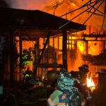 Firefighters in gear battle a large house fire at night, surrounded by flames, smoke, and debris as parts of the structure collapse and burn intensely in the background.