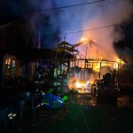 Firefighters work at night to put out a large fire engulfing a building, with smoke and flames rising from the roof. Emergency equipment and hoses are visible in the foreground.