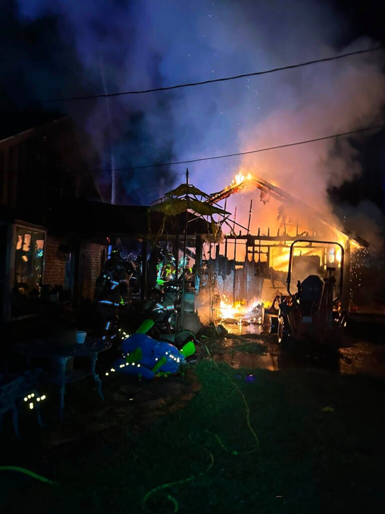 Firefighters work at night to put out a large fire engulfing a building, with smoke and flames rising from the roof. Emergency equipment and hoses are visible in the foreground.